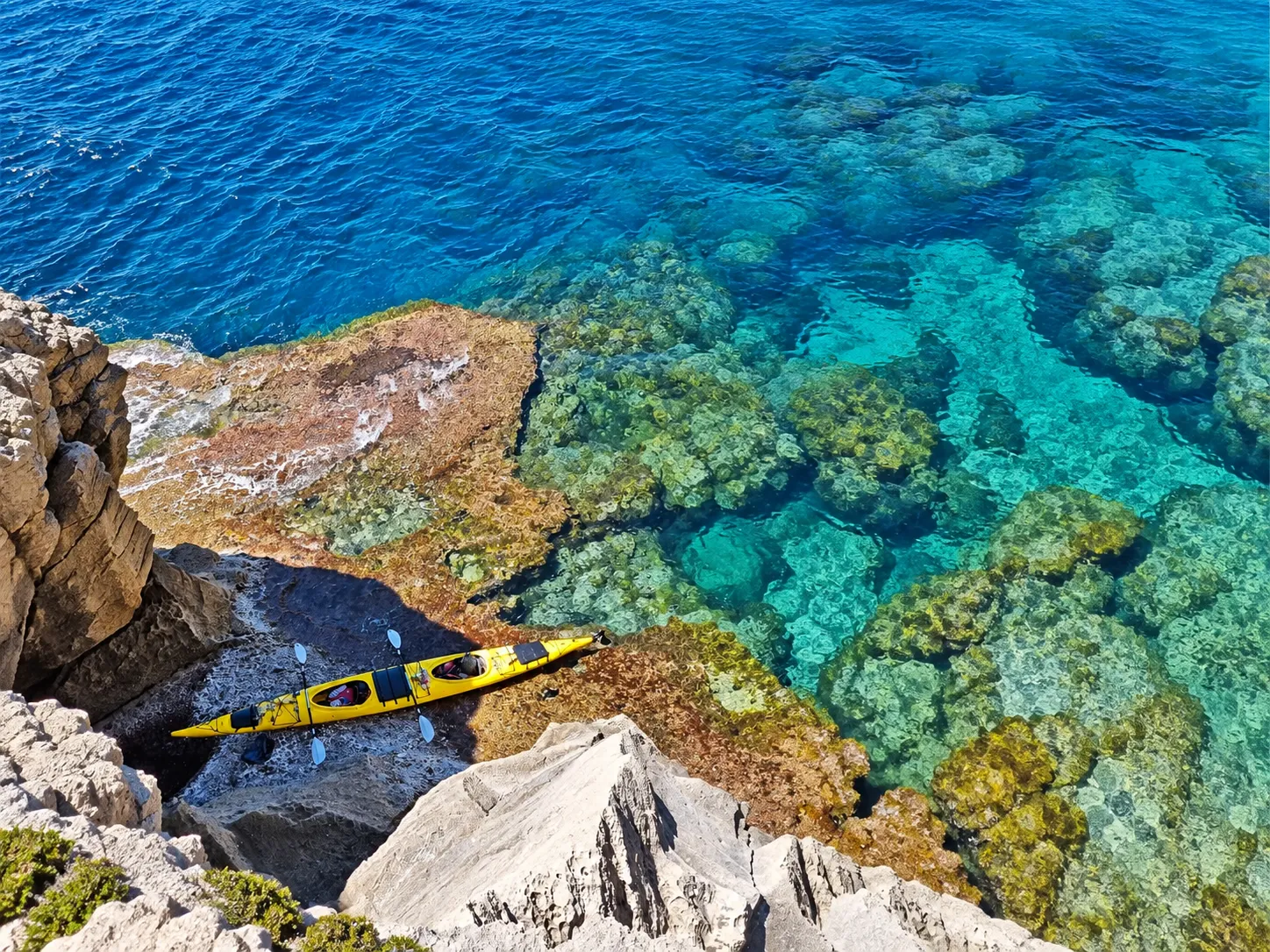 Un kayak de mer double jaune et ses pagaies sur les rochers près d’une eau limpide.